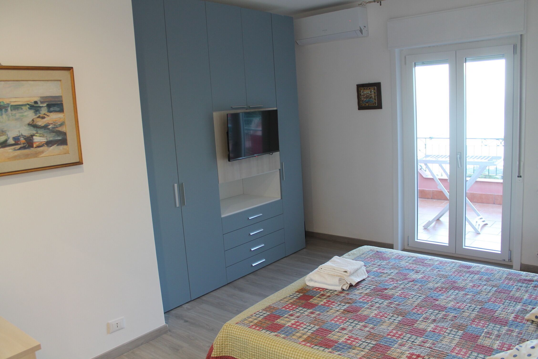 A bedroom with a bed, a TV mounted on a blue wardrobe, a balcony door, and a painting on the wall.