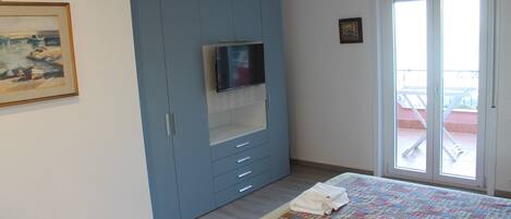 A bedroom with a bed, a TV mounted on a blue wardrobe, a balcony door, and a painting on the wall.