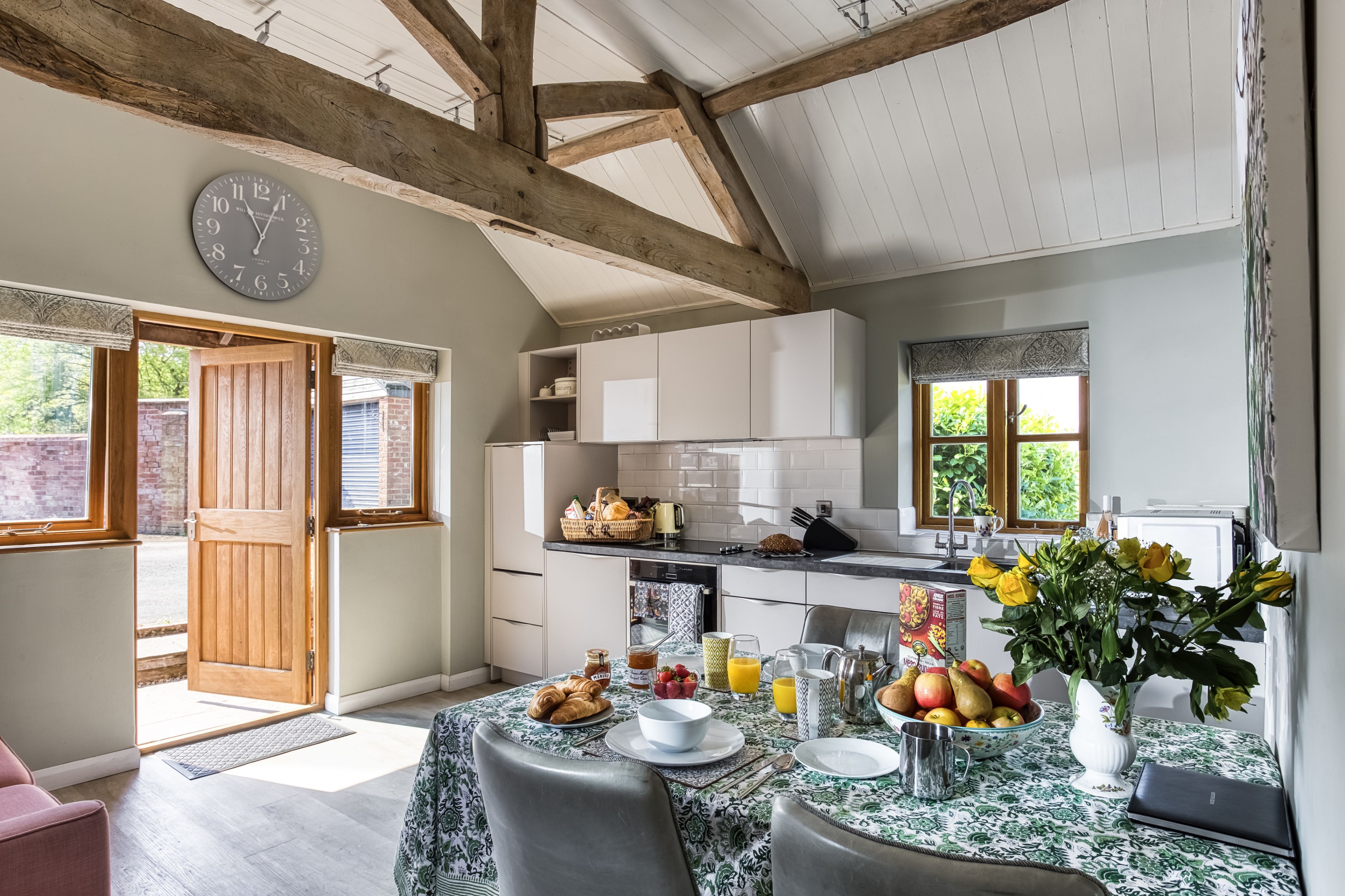 A kitchen with a dining area, wooden beams, a clock, and a window with a view.