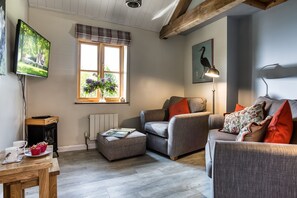 A cozy living room with a grey sofa, a small ottoman, a television, and a window with a view of greenery. - Little Owls Barn, Preston Wynne, Herefordshire (Preston Wynne)