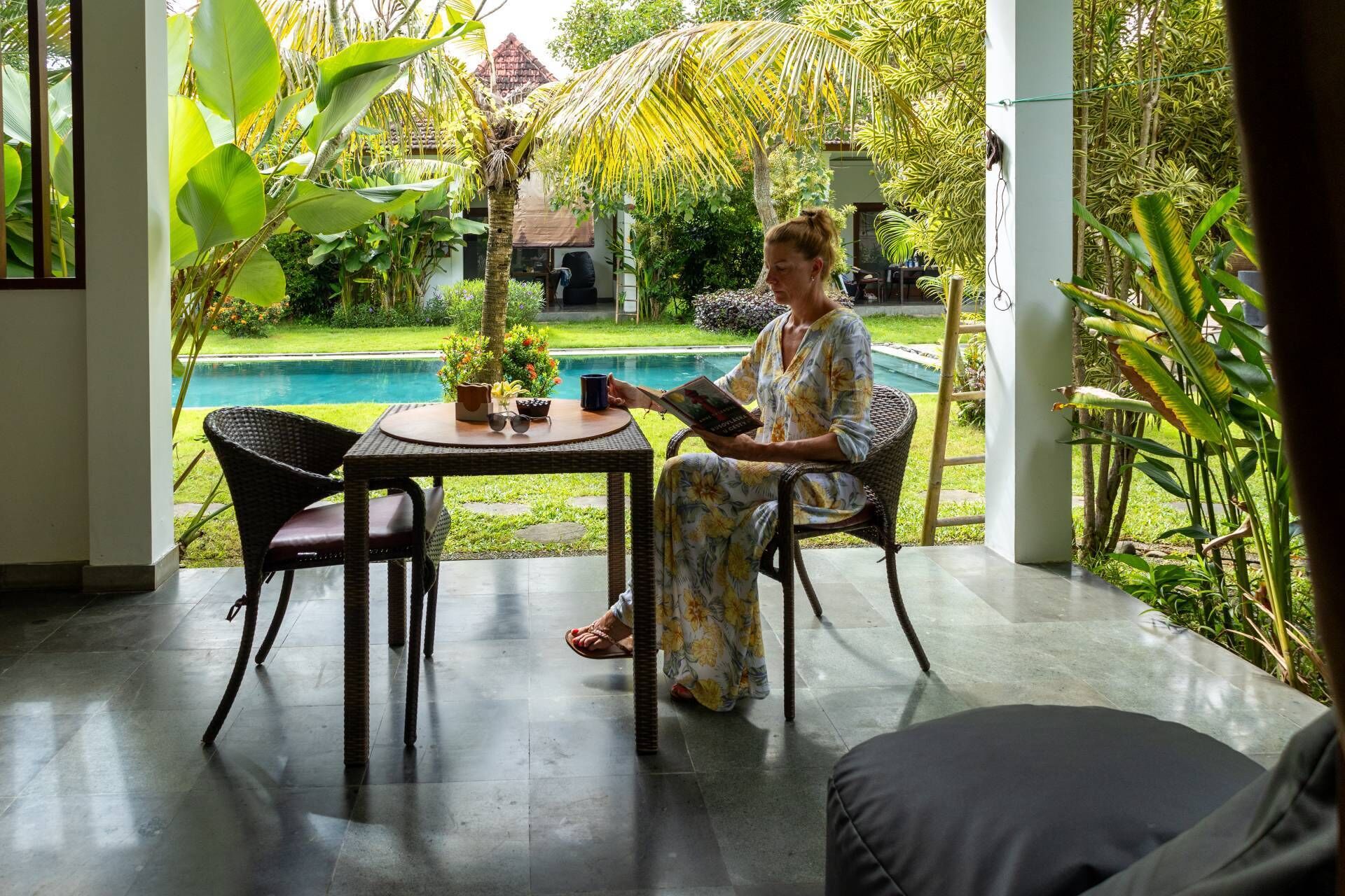 A person seated at a table with a book, in a room with a view of a swimming pool and lush greenery.