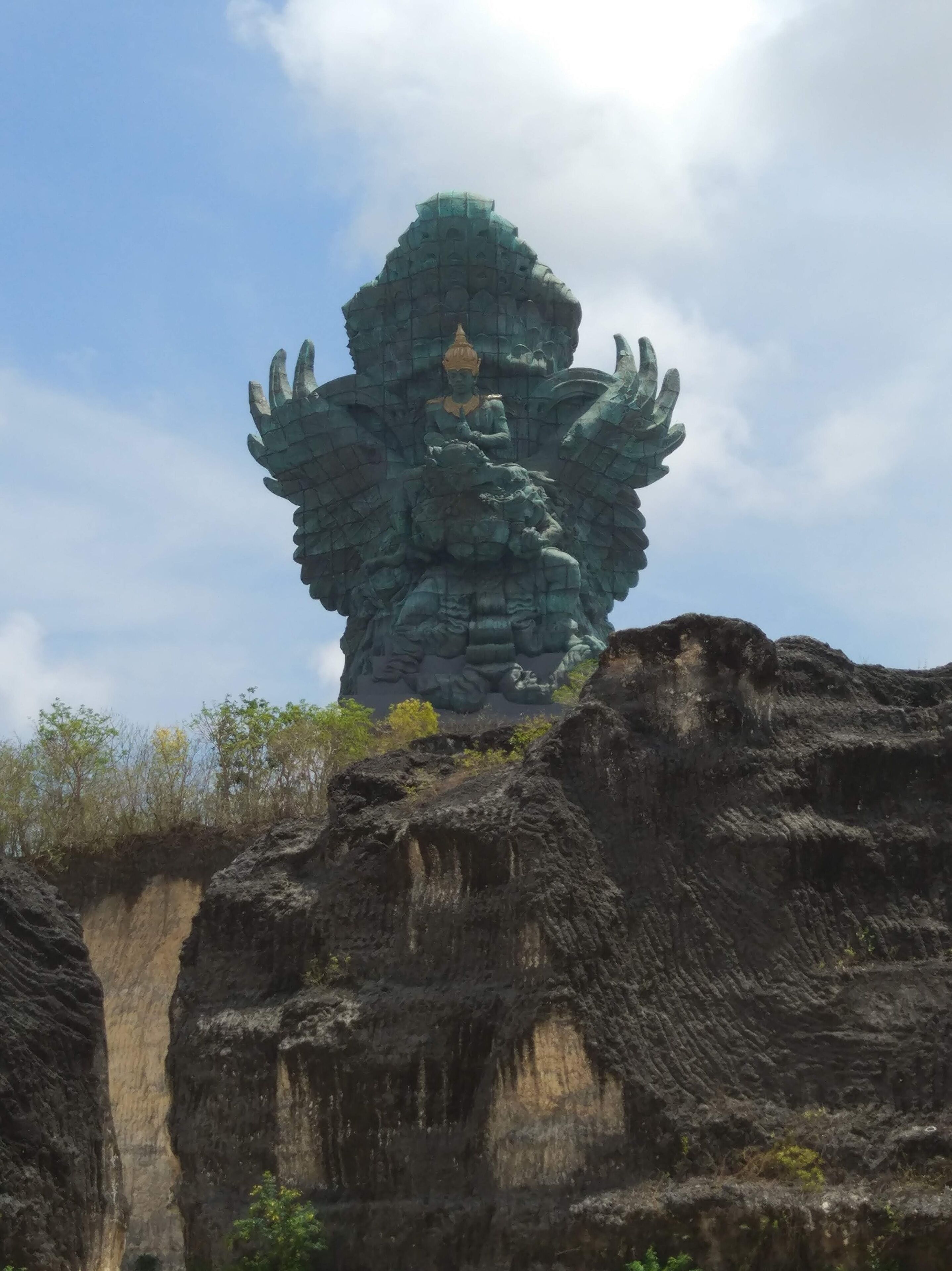 Une grande formation rocheuse sur laquelle se trouve une statue représentant une figure assise sur une fleur de lotus, le tout étant entouré de feuillage.