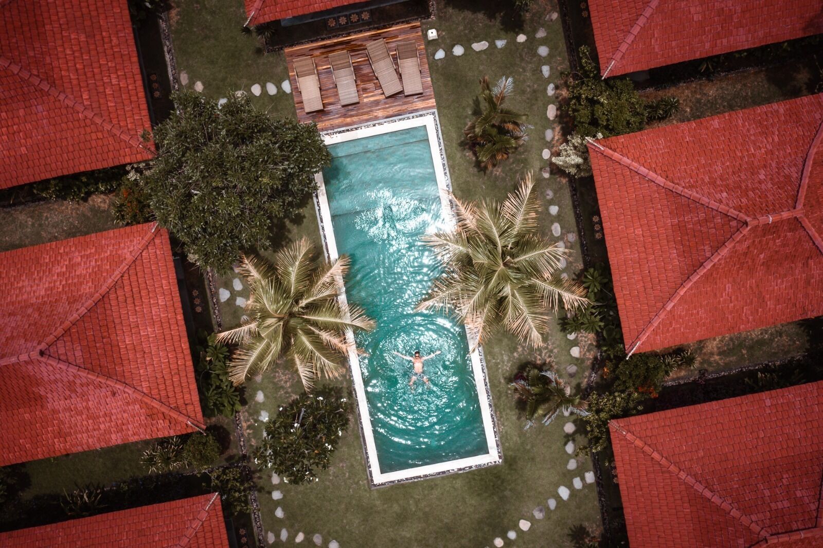 Vista aerea di una piscina rettangolare circondata da palme e edifici con tetti rossi.