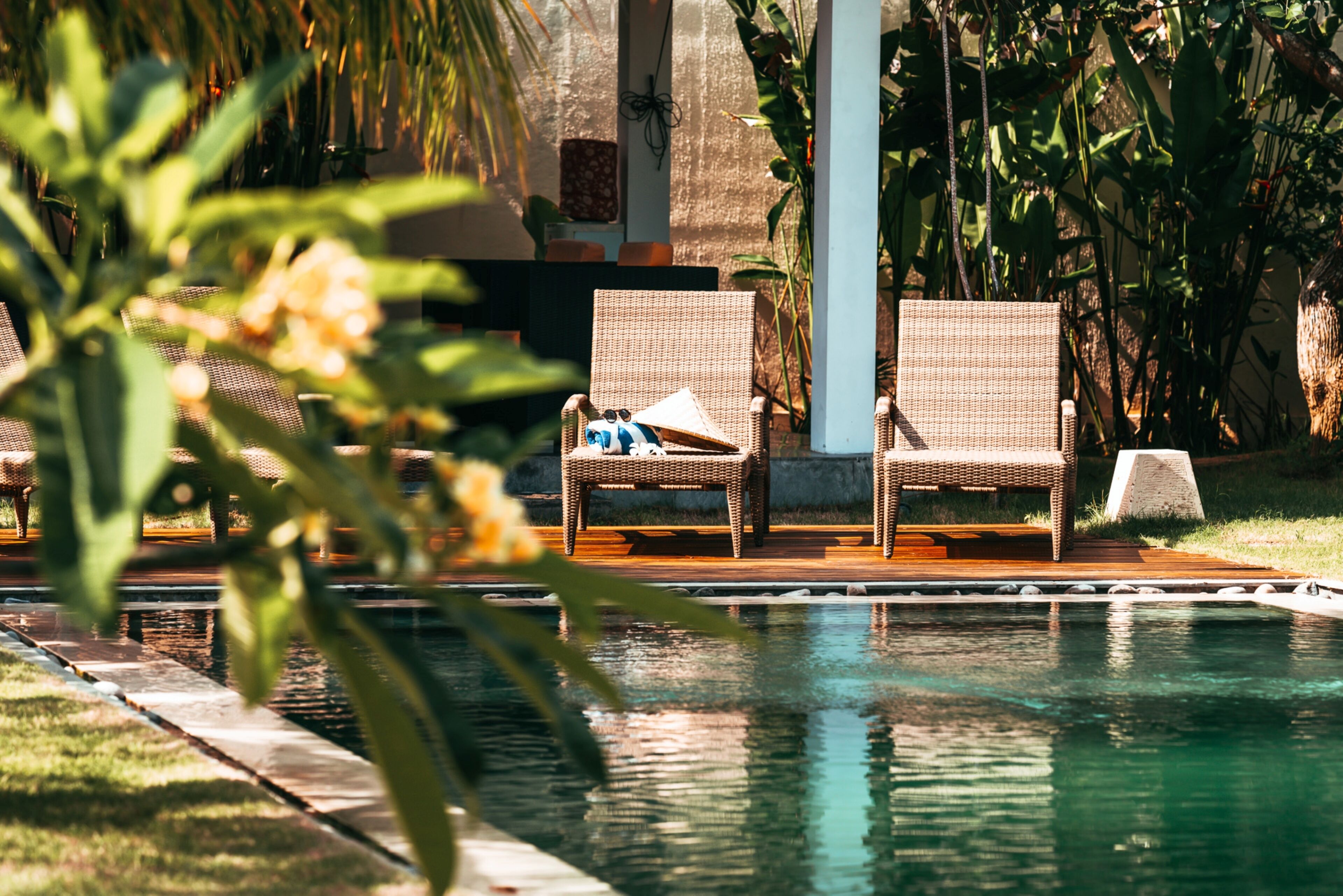 Zona de piscina con sillones de mimbre y una mesa pequeña, rodeada de abundante vegetación.