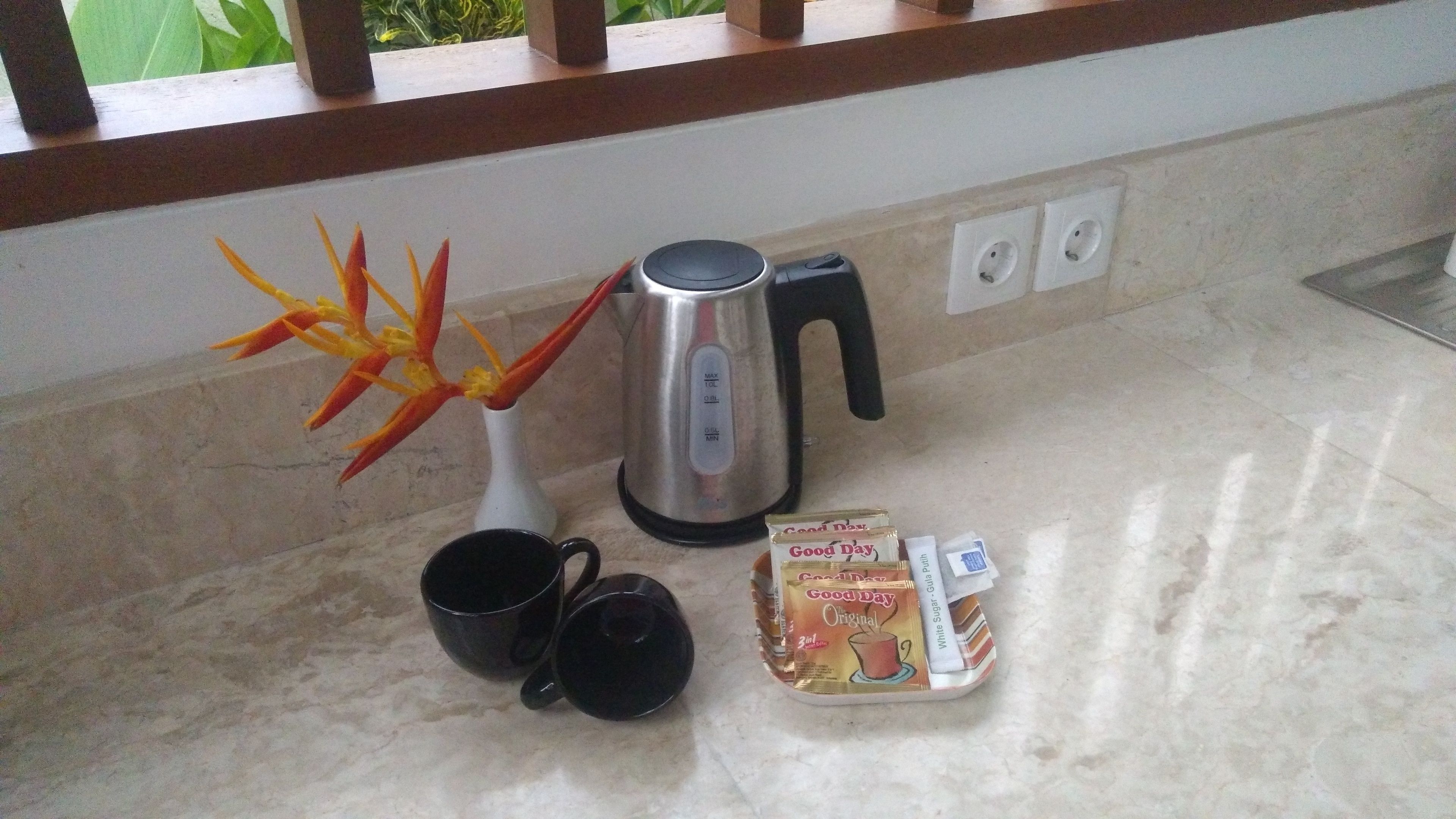 A kitchen counter with a coffee maker, two black mugs, a packet of Good Day tea, and a decorative plant.
