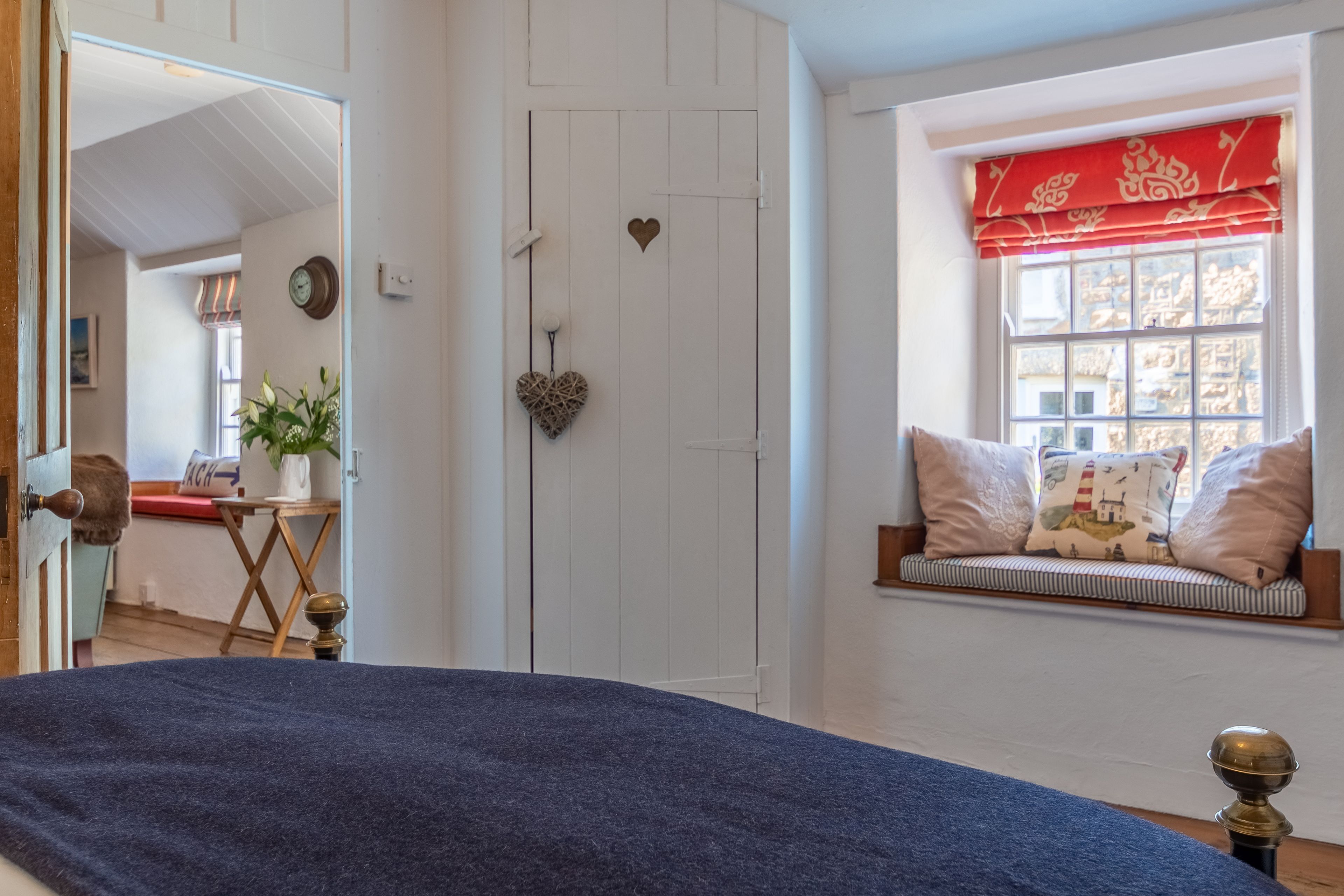 A room with a bed, a window with a red valance, a heart-shaped decoration on the door, and a view of a room with a plant and a table.