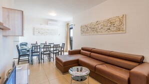 A modern living room with a brown leather sofa, a glass coffee table, and a dining area with a table and chairs. - GetawaysMalta - Seashells 2-bedroom Apartment in Bugibba (San Pawl il-Baħar)