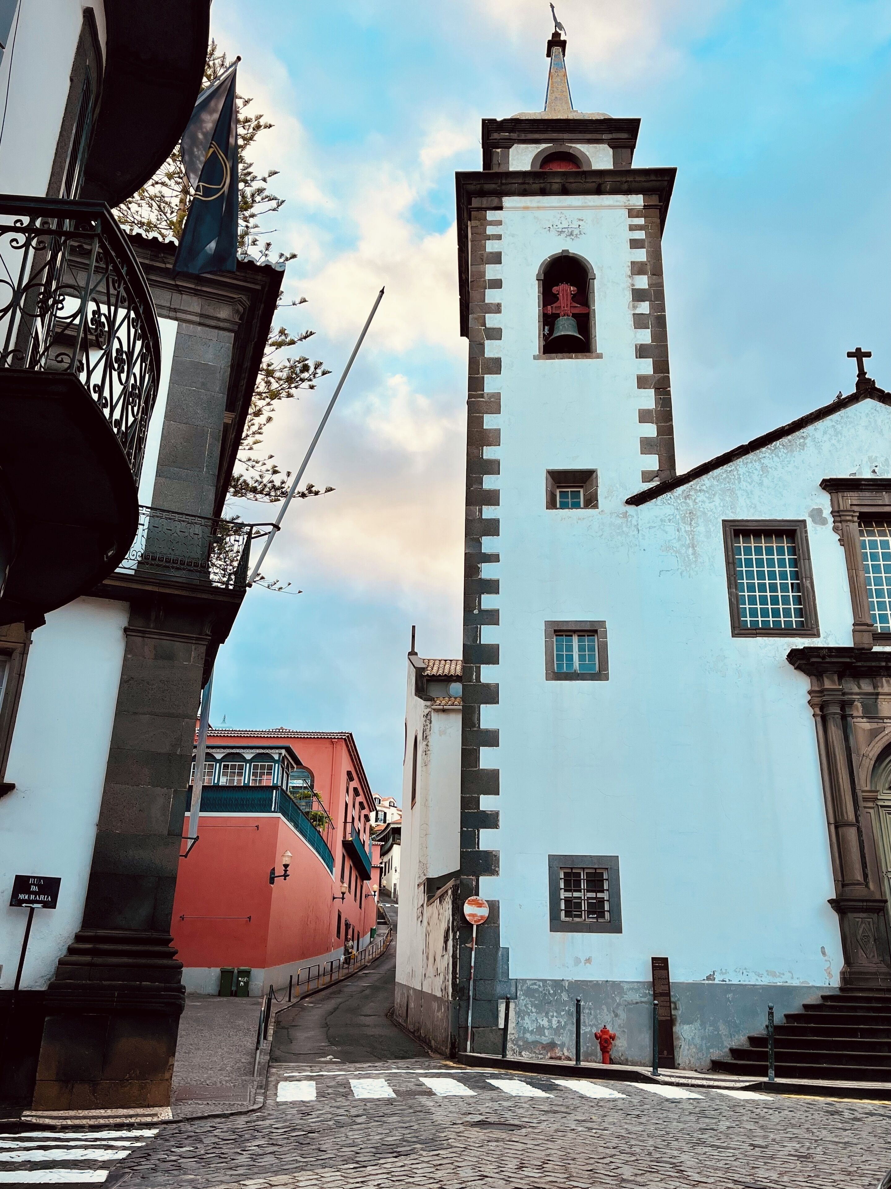 A narrow street with a white building featuring a bell tower and a red building with a balcony.