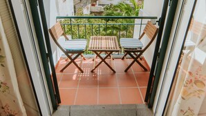 A balcony with two wooden chairs and a small table, surrounded by a glass railing, with a view of greenery and buildings in the background. - Casa da Calçada - 2nd Floor (Funchal)