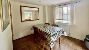 Een eethoek met een tafel bedekt met een tafelkleed met stippen, houten stoelen, een spiegel aan de muur, een raam met gordijnen, en een radiator onder het raam.