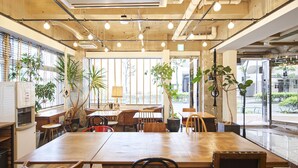 Ruime, overdekte ruimte met een grote houten tafel en meerdere stoelen, omgeven door potplanten en grote ramen met uitzicht op buiten.