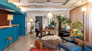 Een moderne hotel lobby met een blauwe accentmuur, een receptiebalie, een eetgedeelte en een zithoek met een bont plaid en blauwe poefs.