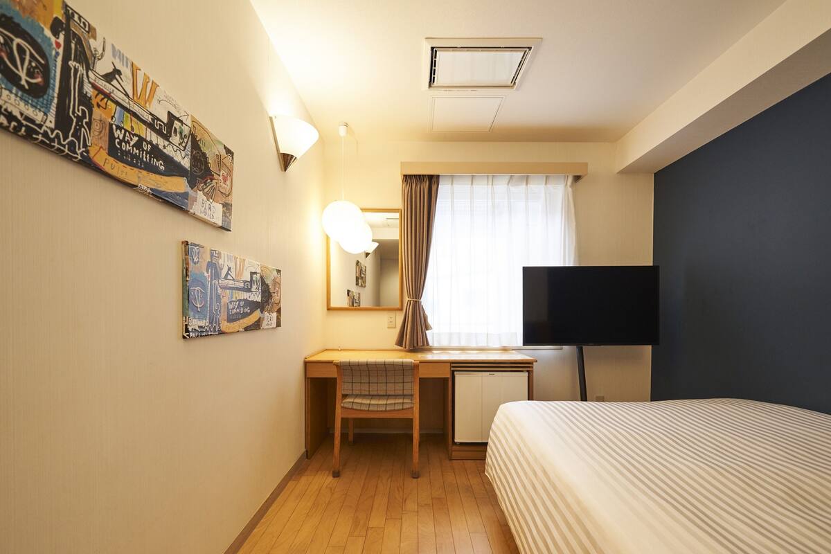 Una habitación de hotel con cama, escritorio, silla, televisor y obras de arte en las paredes.