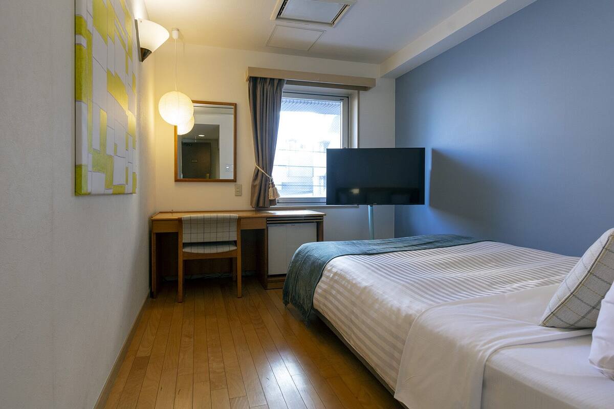 Habitación de hotel con cama, escritorio, silla, televisión y ventana con cortinas.