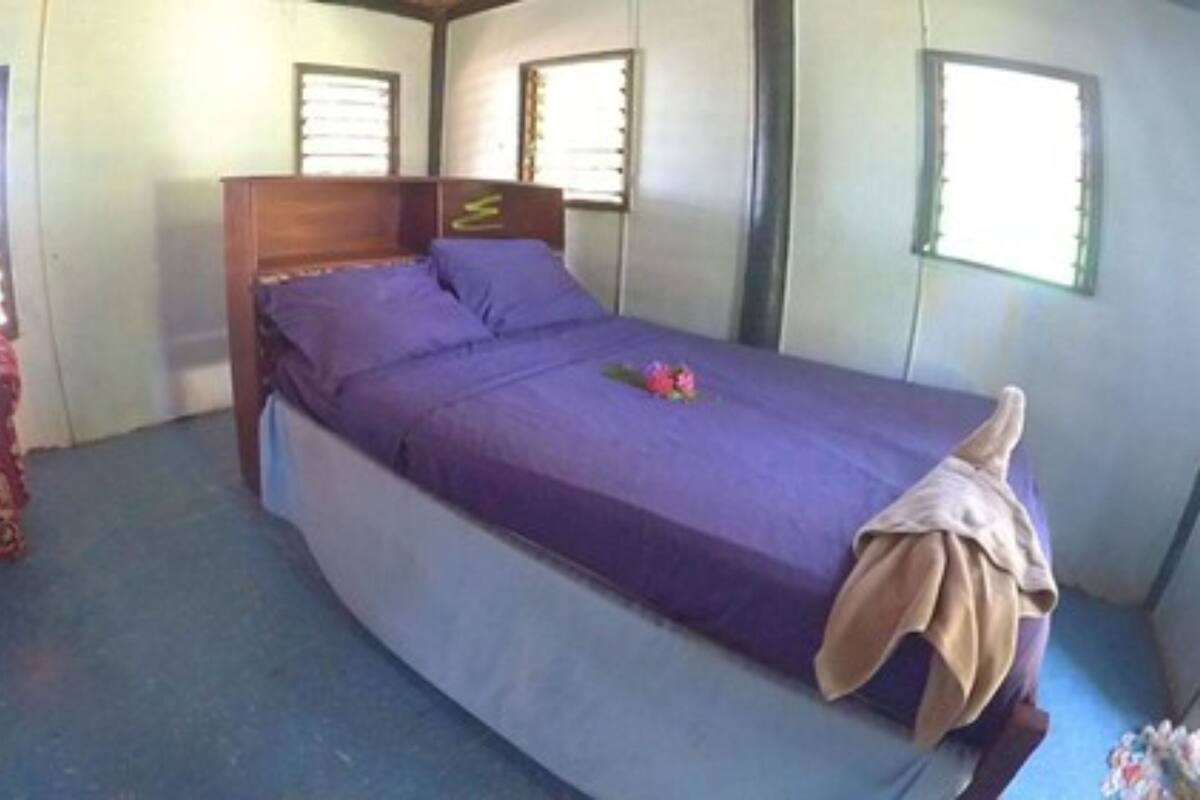 Habitación con una cama individual, cabecera de madera, colcha morada y una decoración floral. Se observa una silla roja con estampado y una mesita con un jarrón de flores.