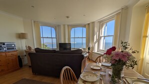 Un coin repas avec une table dressée pour deux personnes, un canapé et une vue sur la mer grâce à de grandes fenêtres.