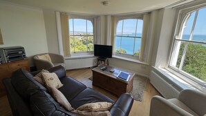 Un salon avec un canapé en cuir, une table basse en bois et un téléviseur. De grandes fenêtres offrent une vue sur la mer et la verdure.