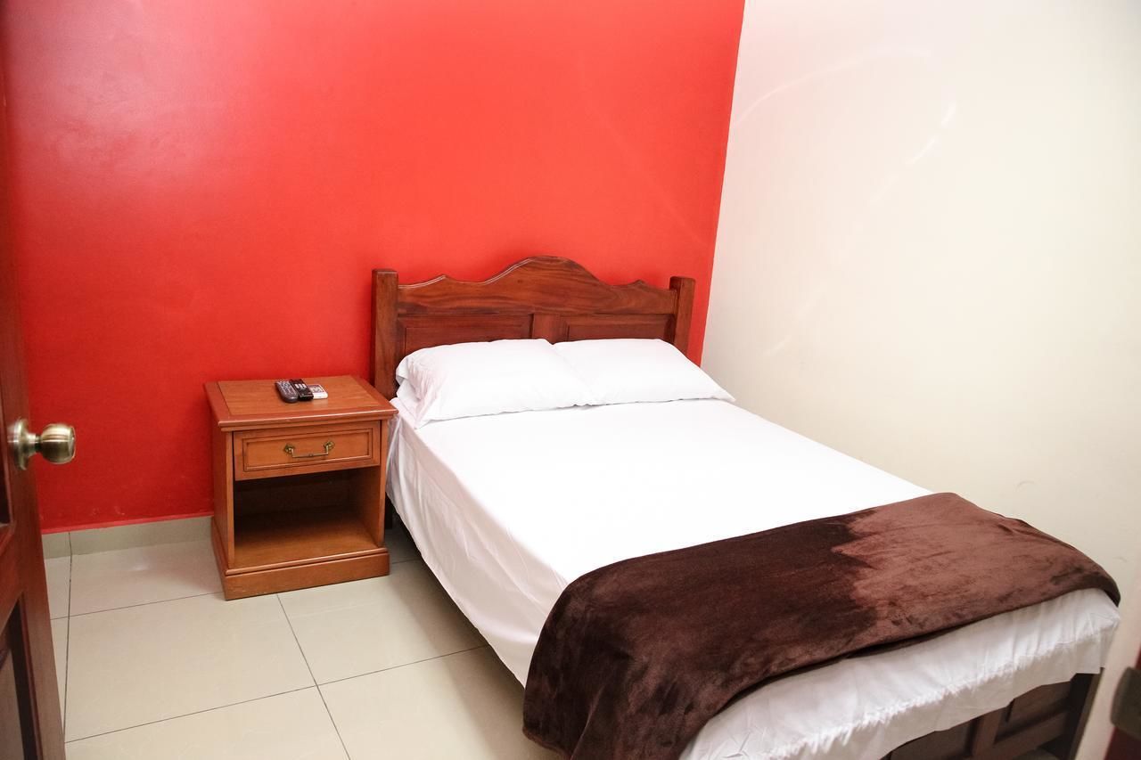 Un dormitorio pequeño y ordenado con una cama de madera, ropa de cama blanca, una manta marrón y una mesita de noche con un control remoto.