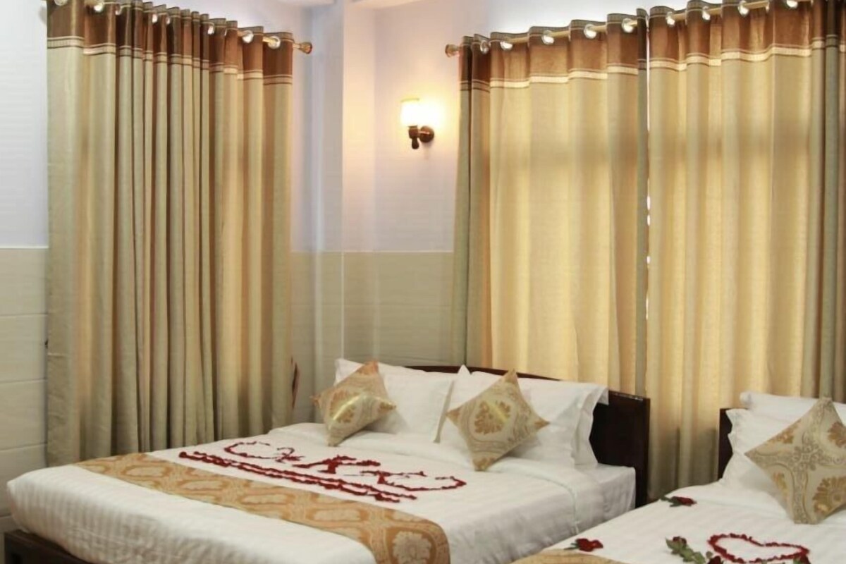 A hotel room with two beds, beige curtains, a wall-mounted light, and a patterned bedspread.