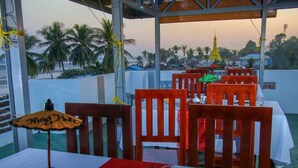 A dining area with wooden chairs and tables, a table umbrella, salt and pepper shakers, a glass of orange juice, and a view of palm trees and a temple in the background. - King Hotel (Katha)