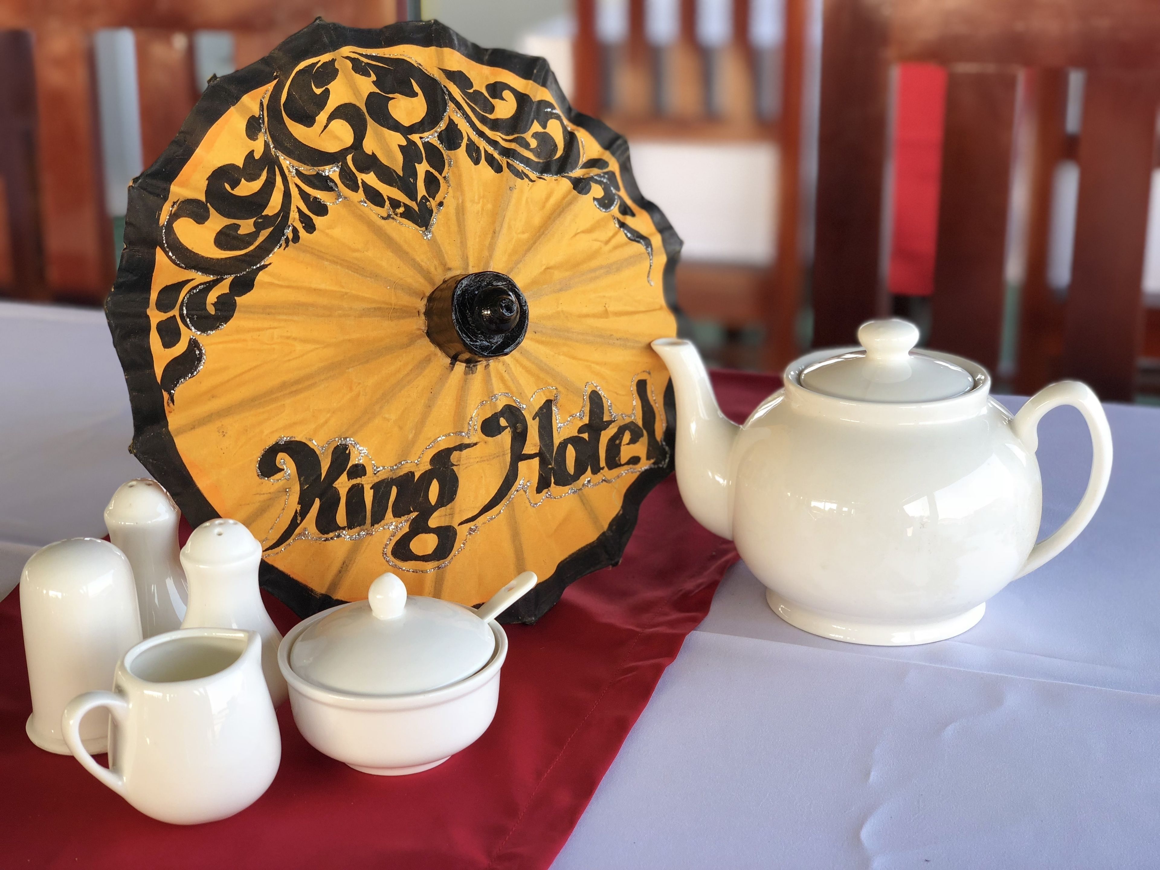 Una mesa dispuesta con un hervidor blanco, terrones de azúcar, una jarrita de leche, una pequeña jarra y un paraguas decorativo con el texto "King Hotel".