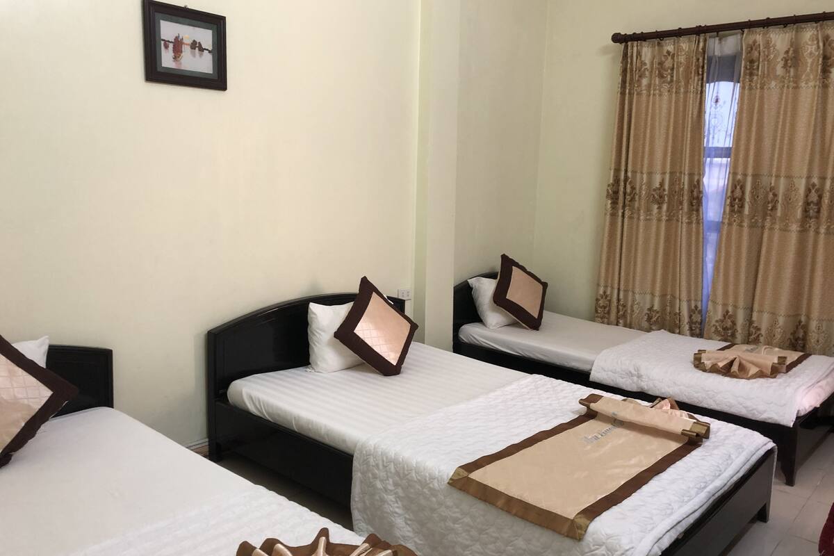 Habitación de hotel con tres camas individuales, cada una con almohada y manta. Hay un cuadro en la pared y una ventana con cortinas.
