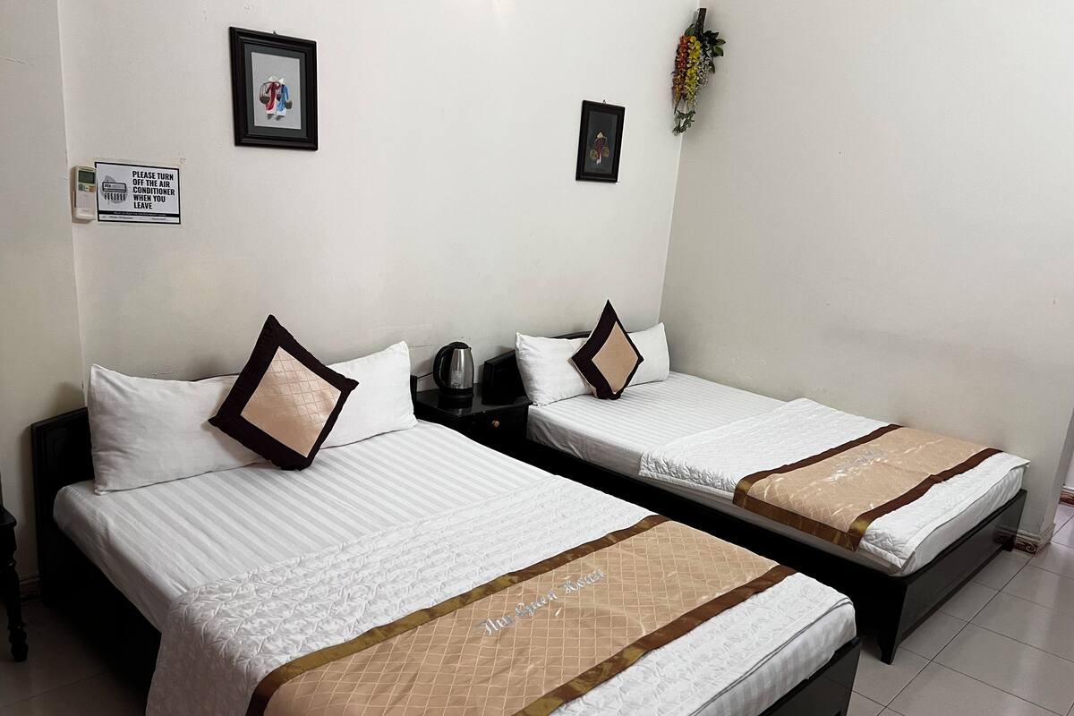 Dos camas individuales con ropa de cama blanca y cabeceros marrones, una mesita con hervidor, y aire acondicionado instalado en la pared.
