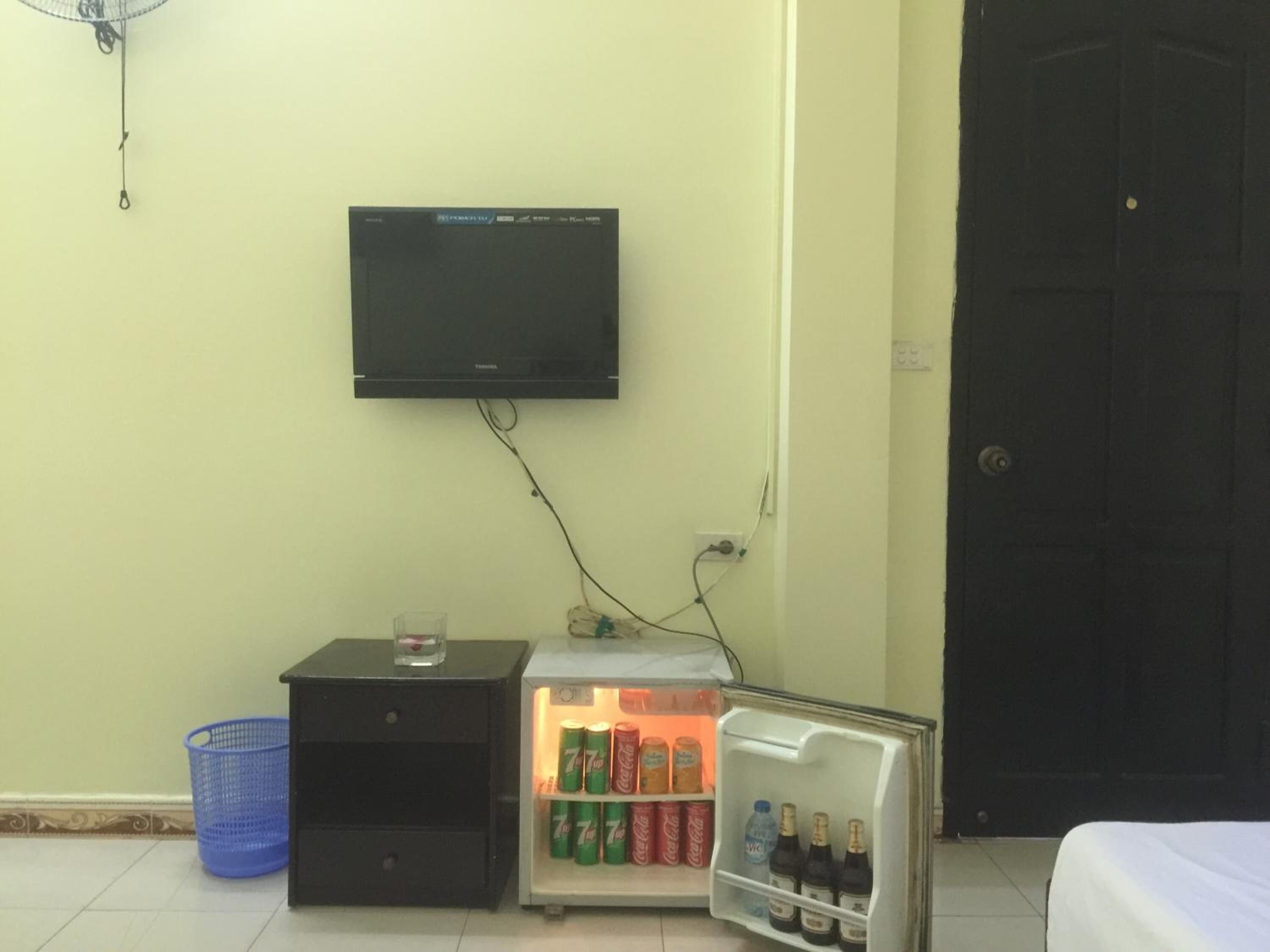 Una stanza con una televisione a parete, un piccolo frigorifero con bevande, un comodino e un cesto blu.