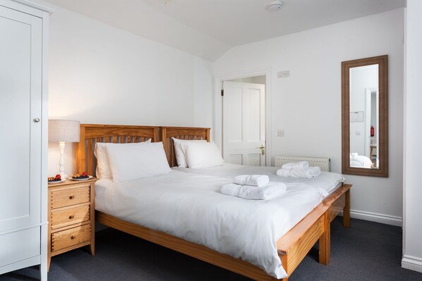 A neatly made bed with white linens and towels, a bedside table with a lamp and fruit, and a large mirror on the wall.