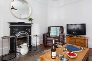 A living room with a fireplace, a leather armchair, a television, and a table with a bottle of wine and strawberries.