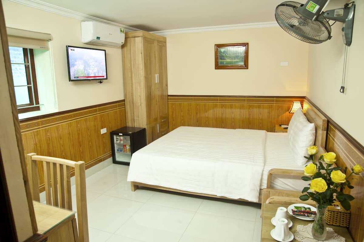 Ein Hotelzimmer mit einem Bett, einem Fernseher, einem Ventilator, einem Holzschrank, einem Stuhl und einem Tisch mit Blumen in einer Vase.