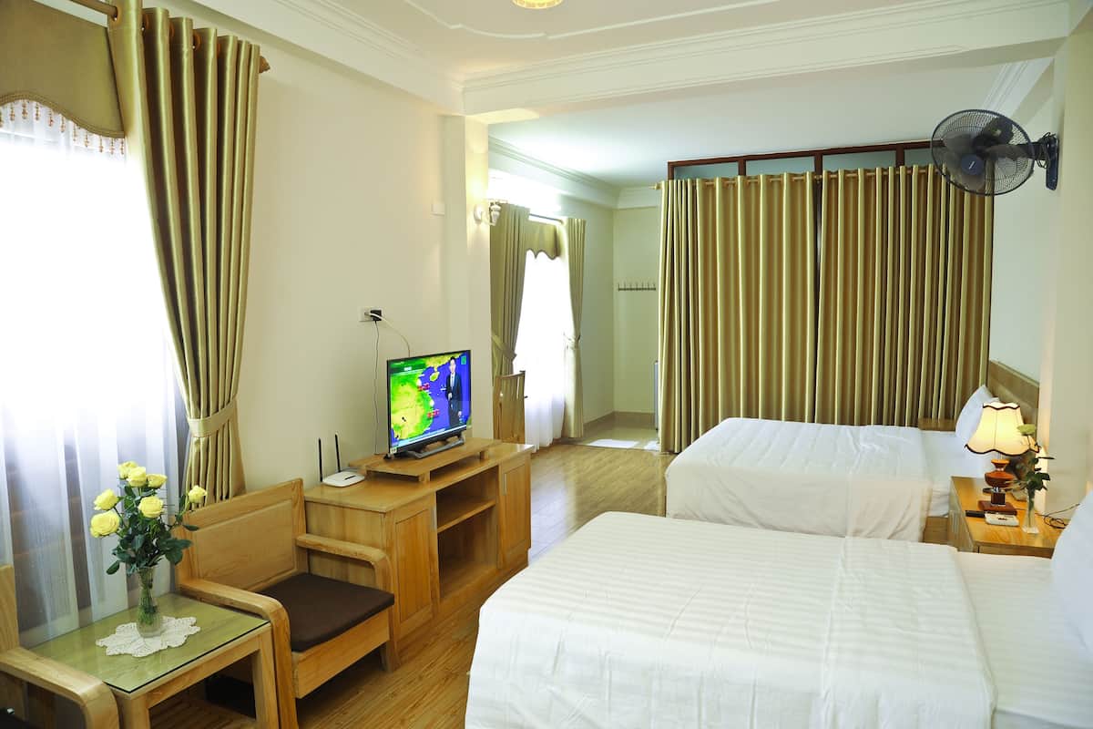 Ein Hotelzimmer mit zwei Betten, einem Fernseher auf einem Holzgestell, einem Ventilator und einem kleinen Tisch mit einer Blumenvase.