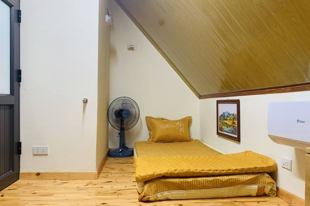 Ein kleines Zimmer mit einem Bett, einem Standventilator, einem kleinen Schreibtisch und einer Wand-Klimaanlage.