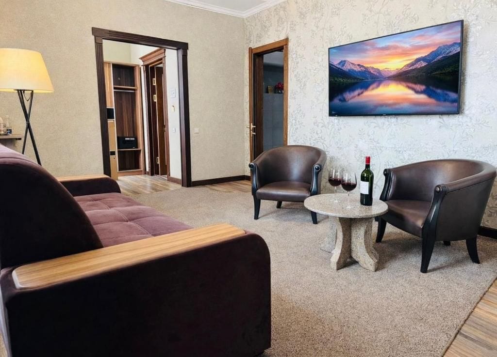 A room with a sofa, two armchairs, a small round table with a bottle and glasses, a lamp, and a painting of a mountain landscape.