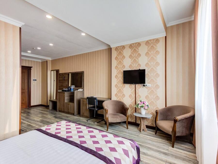 A hotel room with a bed, two armchairs, a desk with a chair, a TV, and a small table with a vase.