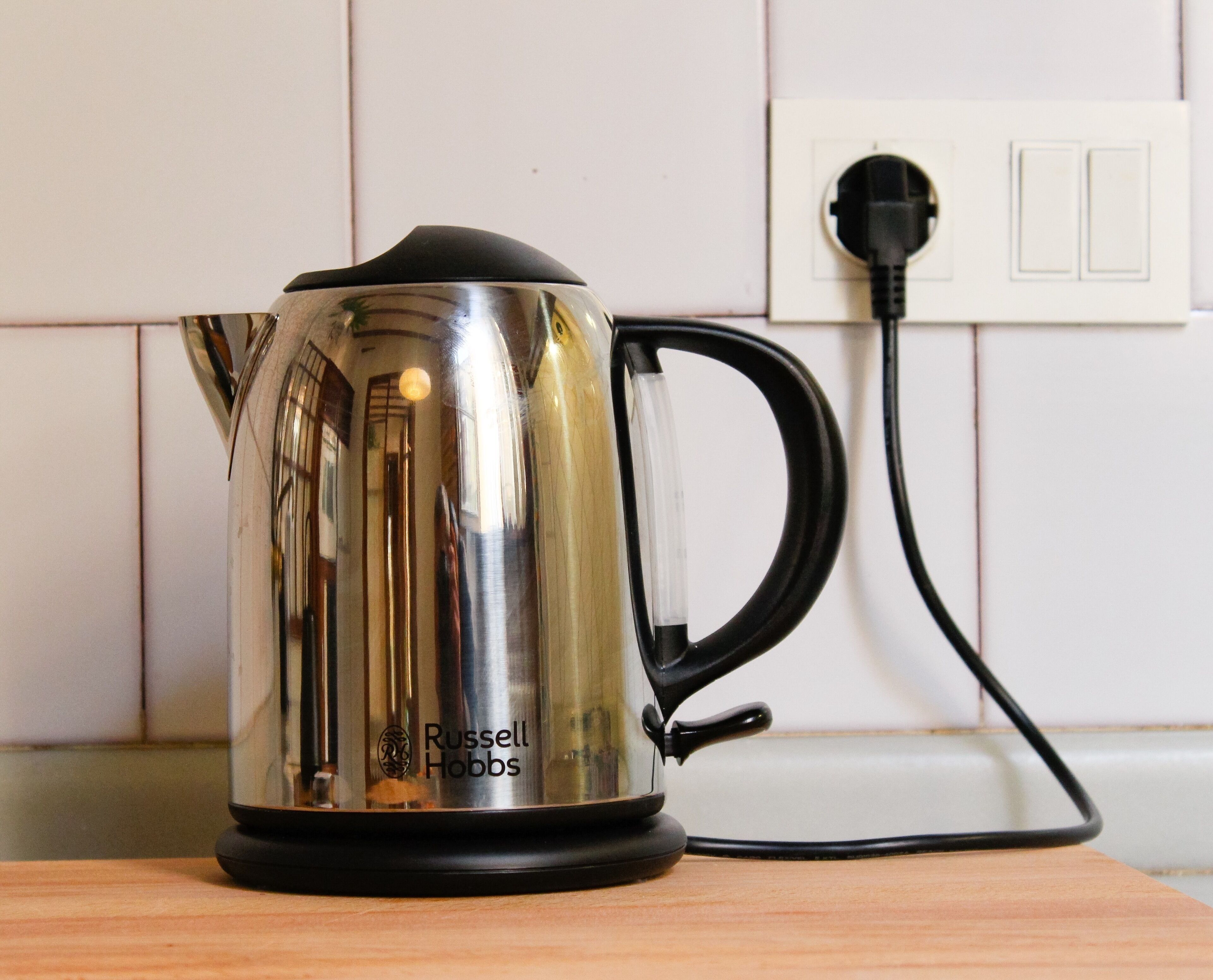 Une bouilloire en acier inoxydable avec une poignée et un socle noirs, portant la marque « Russell Hobbs », branchée sur une prise électrique blanche située sur une surface en bois.