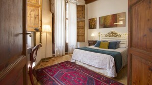 Une chambre avec une porte en bois, un lustre, un lit recouvert d’une courtepointe blanche et verte, un tapis rouge à motifs et des murs en bois.