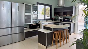 A modern kitchen with stainless steel appliances, a central island with stools, and a large window providing natural light. - 5 Bedroom Villa With Heated Pool, Garden With BBQ & Views Over Hills And Ocean (Adeje)