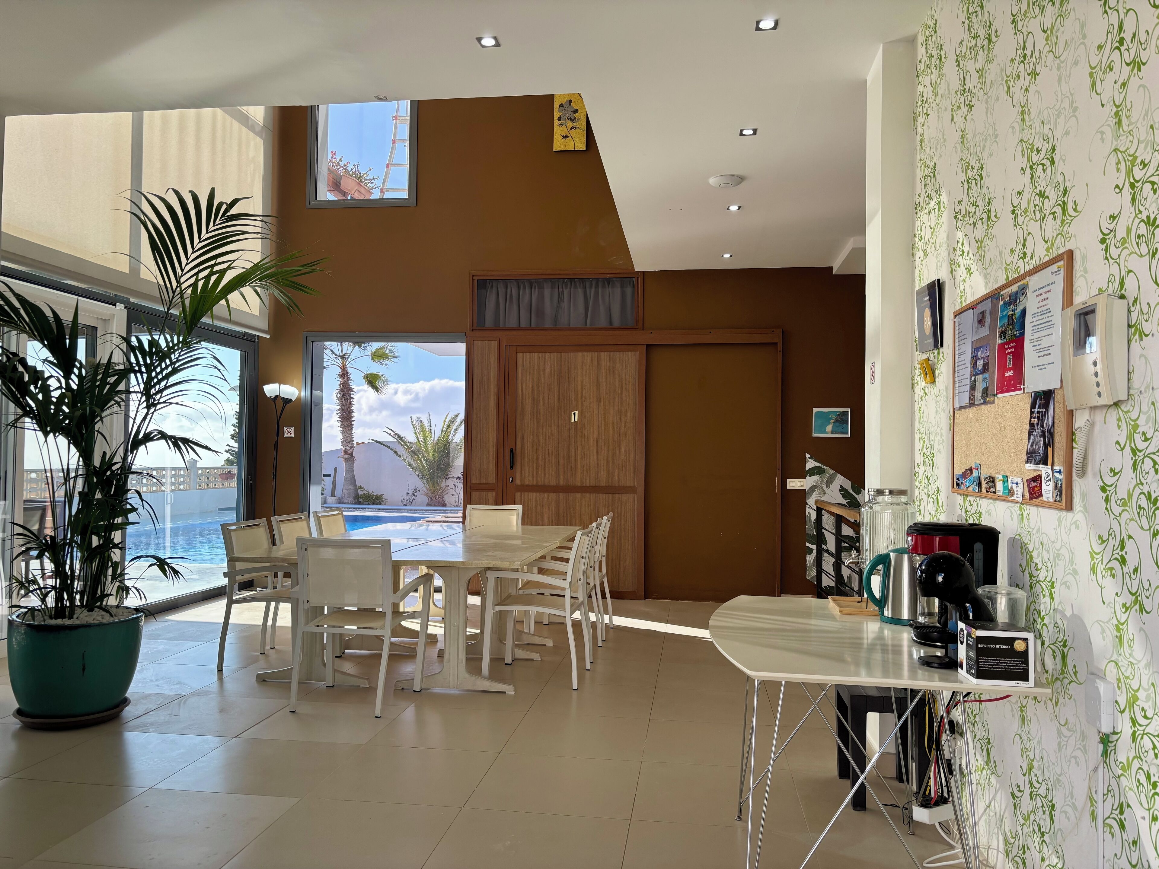 Ein modernes Hotelzimmer mit großem Fenster mit Blick auf einen Pool, einem Essbereich mit weißer Möbel und einer Küchenzeile mit einer Kaffeemaschine.