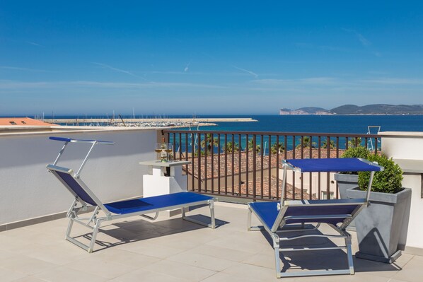 Un balcón con dos tumbonas y una mesa, con vistas a una playa y un puerto deportivo.
