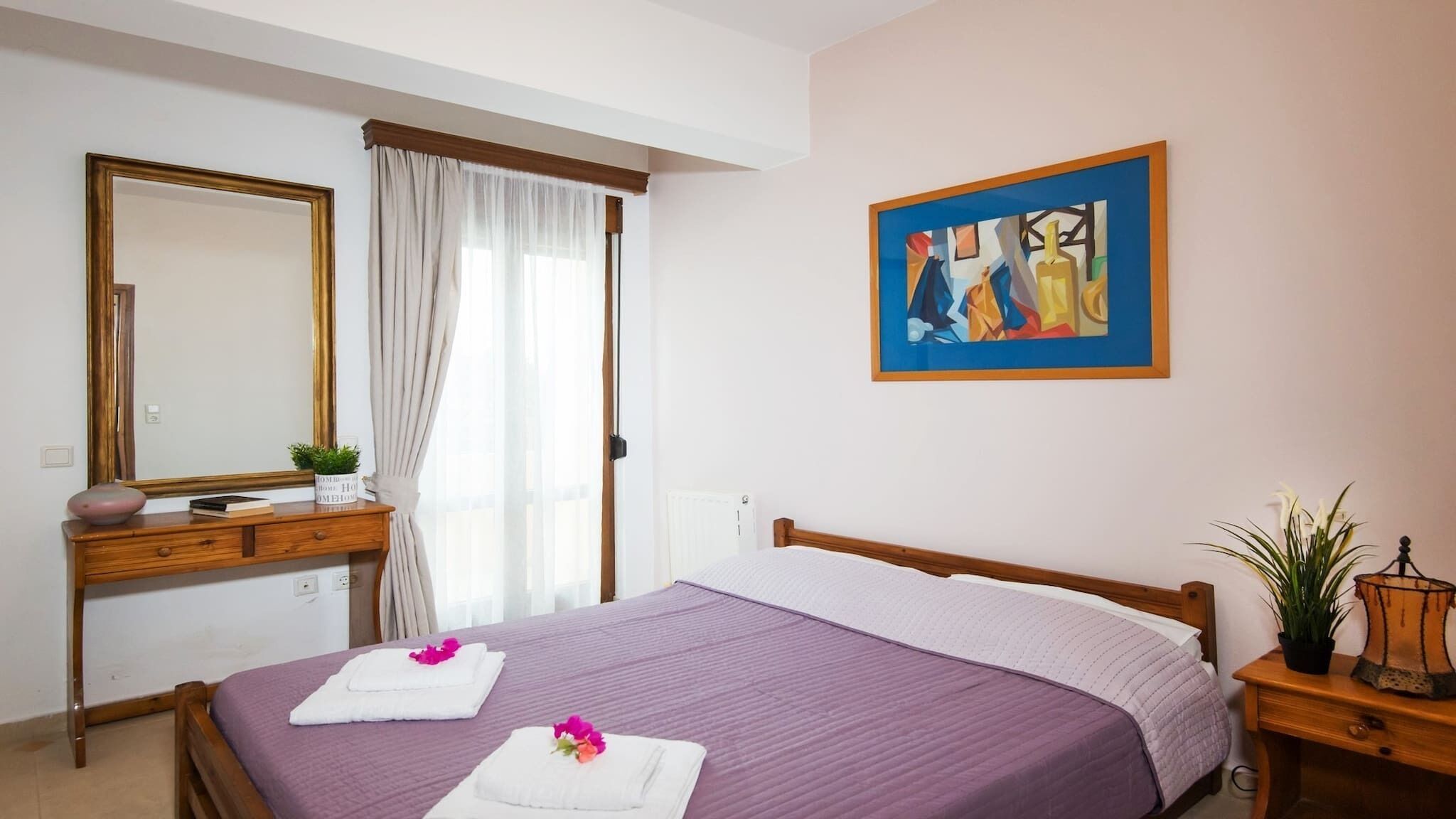 Habitación de hotel con cama, cómoda de madera, espejo, un cuadro y ventana con cortinas.