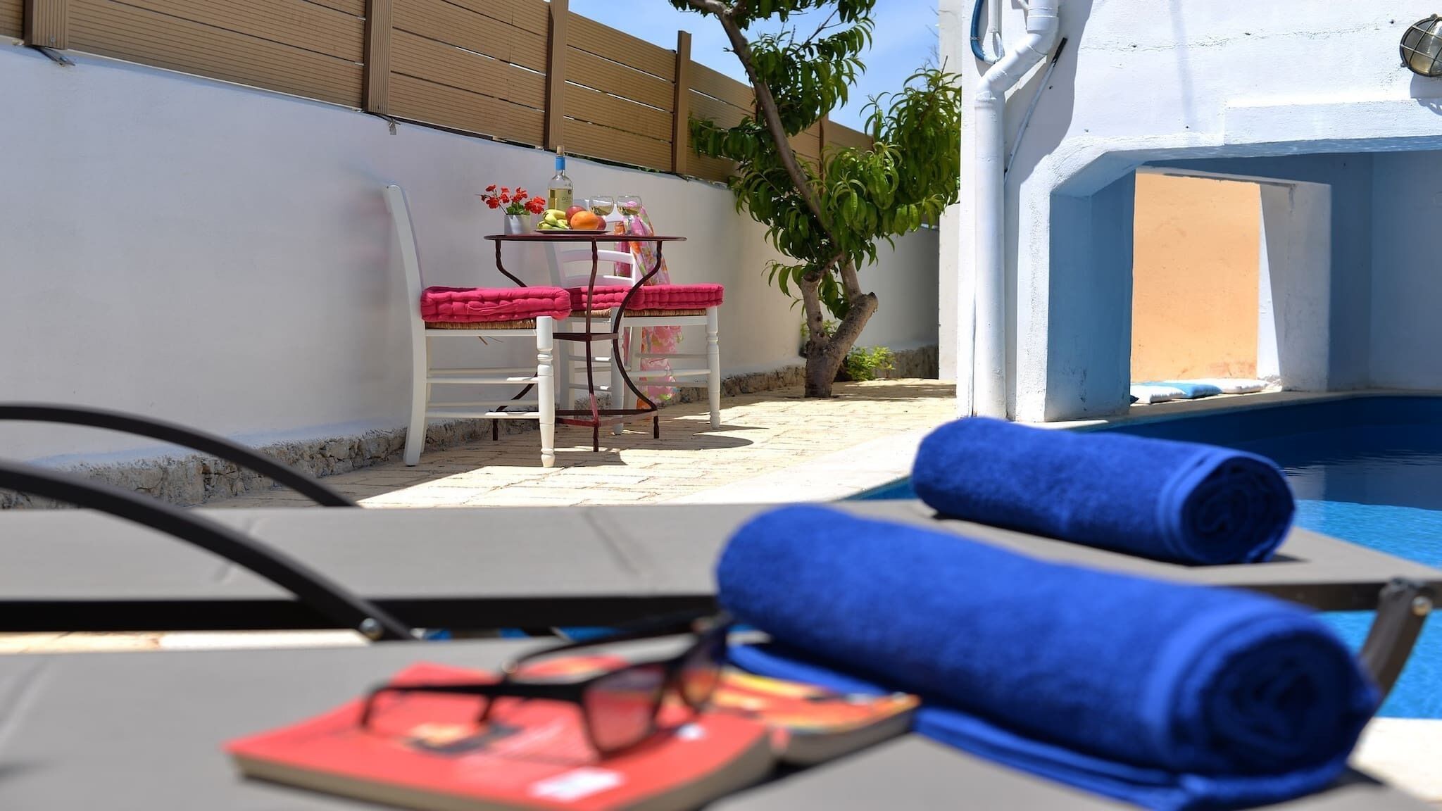 A poolside area with a table set with a bottle, glasses, and fruit. There are two lounge chairs, one with a blue towel and another with a red towel. A pair of flip-flops is on the table.