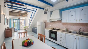 Een keuken met witte kastjes, een gasfornuis en een spoelbak. Er staat een eettafel met stoelen en een schaal vol fruit. Een trap leidt naar een verdieping.