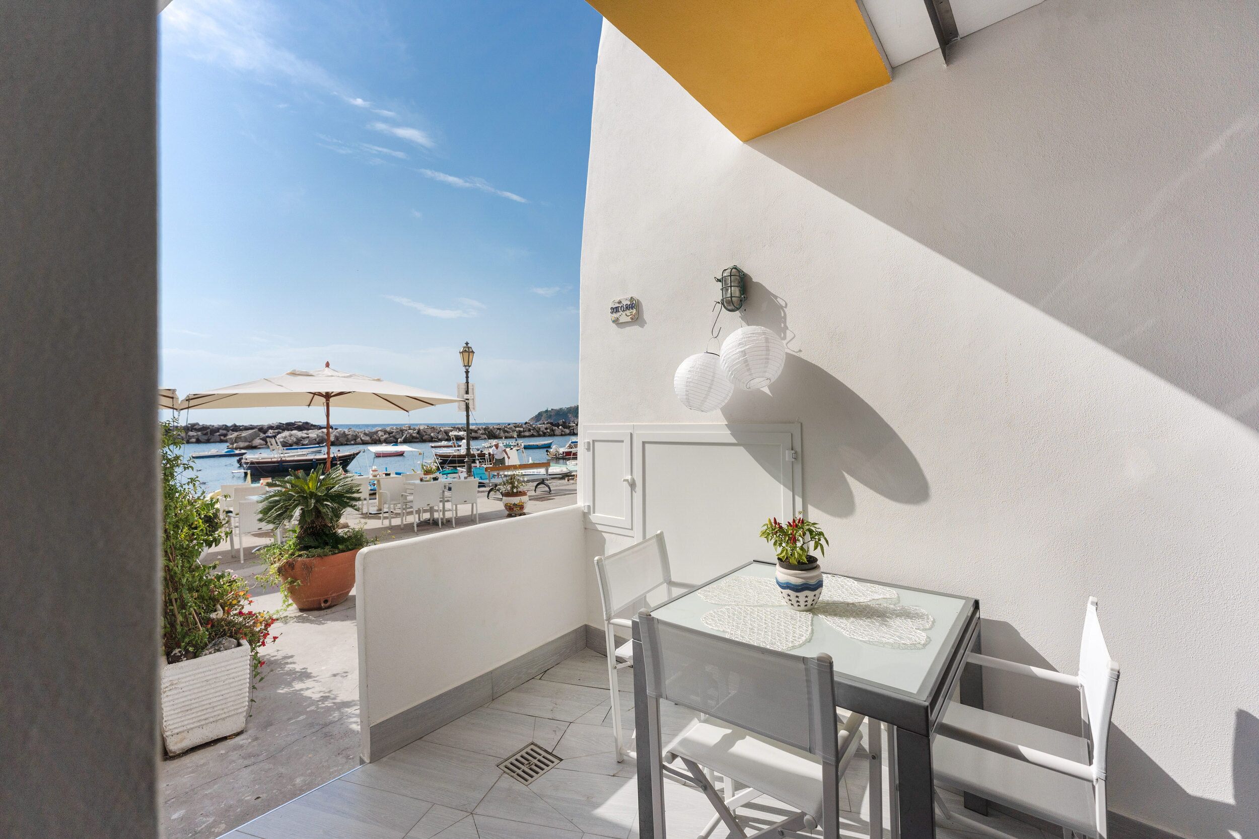 Een balkon met een tafel en stoelen, plantenbakken en uitzicht op zee en boten.