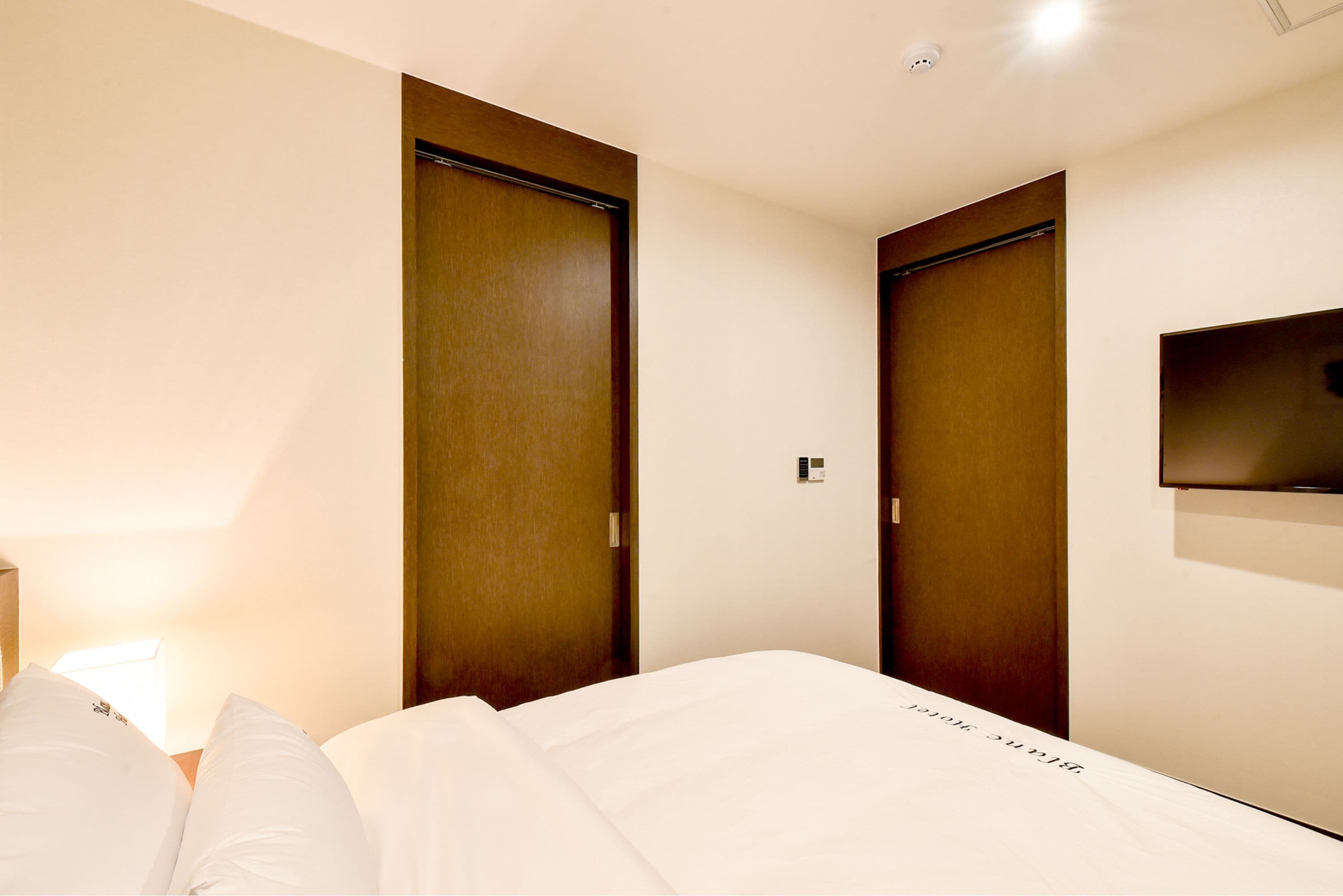 A hotel room with a bed, two wooden doors, a wall-mounted TV, and a bedside lamp.