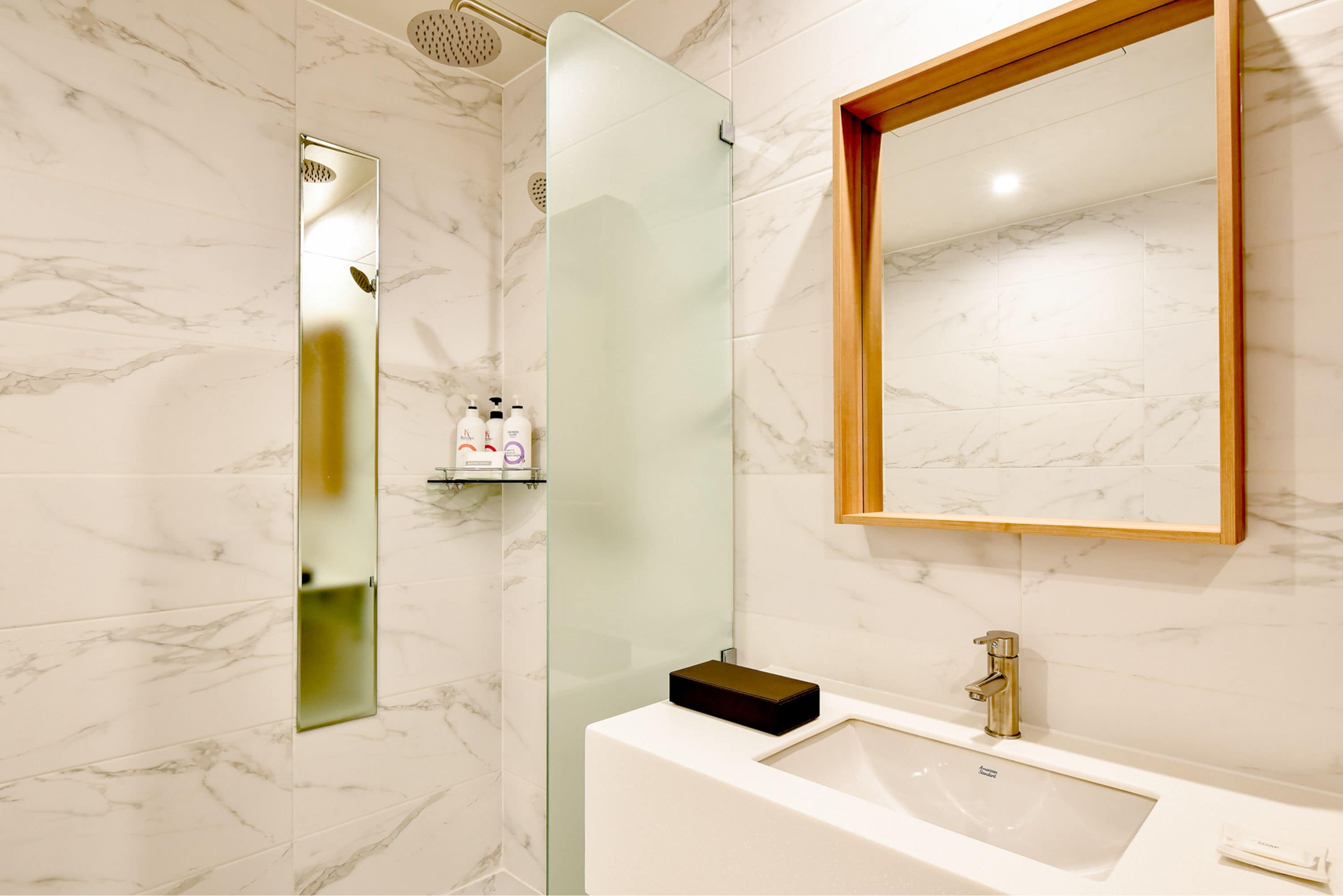 Ein modernes Badezimmer mit Duschkabine aus Glas, Marmorfliesen an der Wand, einem Spiegel mit Holzrahmen und einem weißen Waschbecken mit goldfarbenem Wasserhahn.