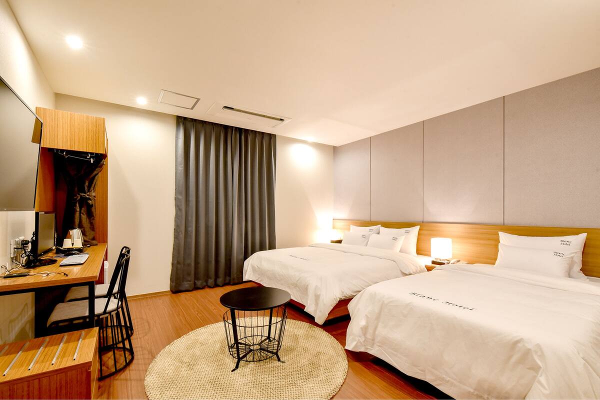 A hotel room with two beds, a desk, a chair, a small table, and a large window with curtains.