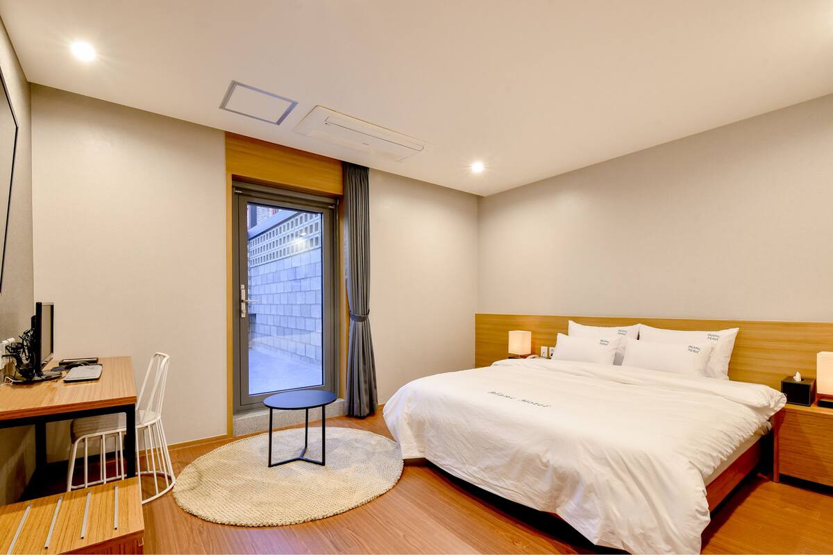 A hotel room with a bed, a desk, a chair, a round rug, and a window with curtains.
