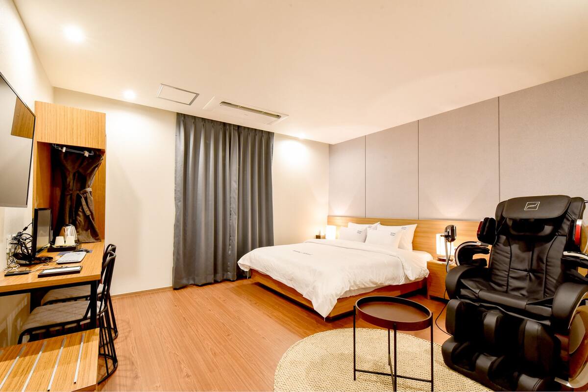 A hotel room with a bed, a massage chair, a desk with a chair, and a small table.