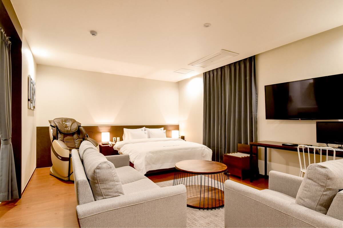 A hotel room with a bed, a massage chair, a sofa, a small table, and a TV.